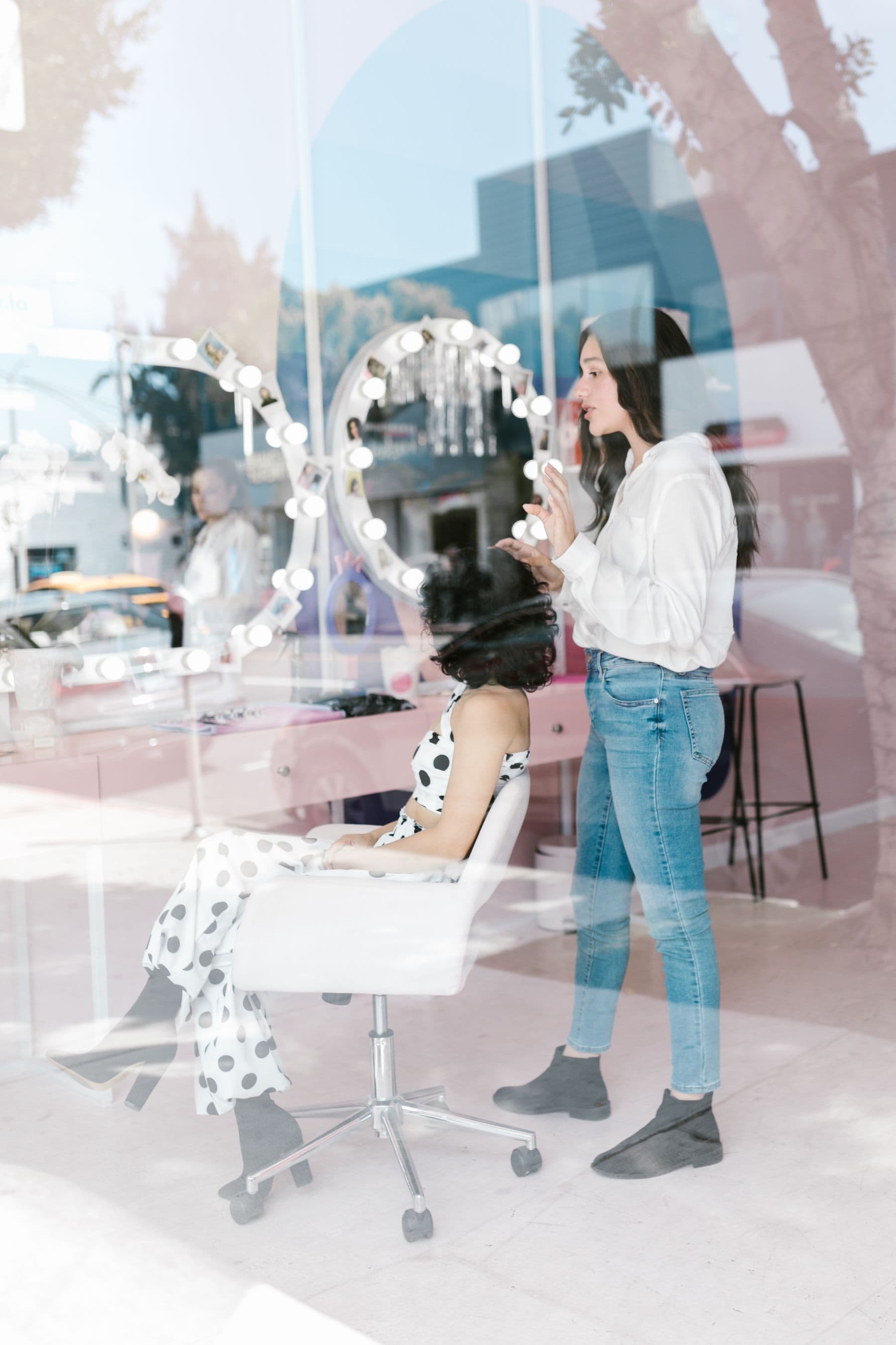 A hair stylist with a seated client, seen through a daylit salon window